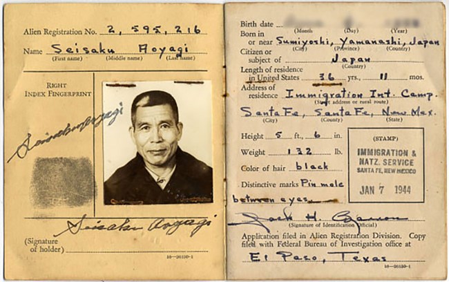 Alien registration card of Seisaku Aoyagi. Collection of Smithsonian National Museum of American History (2015.0252). Pages of a document booklet. On the left, a black and white photo of a Japanese man with short hair, a fingerprint, and a signature. On the right are his identifying characteristics including height, weight, and the presence of a small mole.