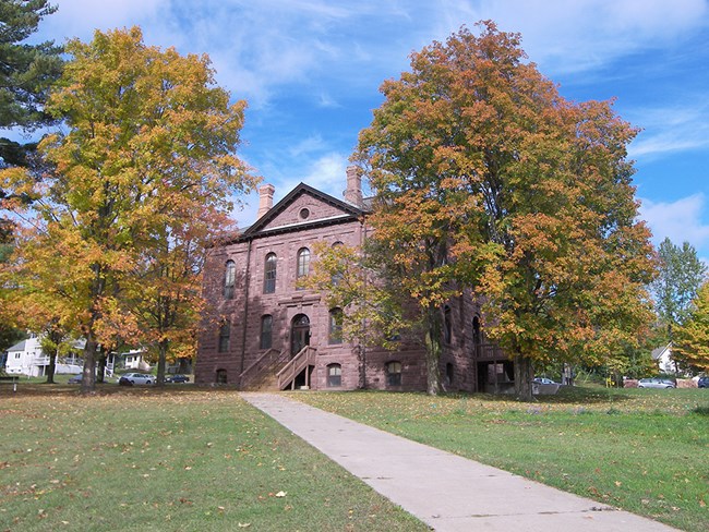Now the headquarters building at Apostle Islands National Lakeshore, this building (the Old Bayfield County Courthouse) was used to house POWs during World War II. Photo by NPS. Color photo. A three-story building of red sandstone sits between two trees just beginning to turn colors in the fall. An area of grass and a sidewalk lead up to the building. The sky is bright blue.