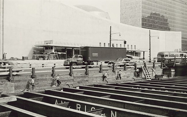 A group of young boys explore exposed steel girders during the construction of the UN Headquarters in NYC. Photo by Angelo Rizzuto, May 1952. Collection of the Library of Congress (https://lccn.loc.gov/2020635609). Black and white photo. Young boys are dwarfed by the size of the steel girders forming supports for an underground area. Behind them, the curved front of the UN building is mostly completed.