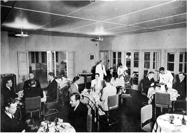 The interior of the Midway Hotel dining room, 1930s. The servers include Chamorro men hired from Guam. Courtesy Pan Am Historical Foundation / paa.org. Several round tables with white table cloths, napkins, and good china fill a room. Many have 2 to 4 people (mostly men) sitting at them. They are being served by men in white uniforms. The lobby of the hotel is visible through an open door.