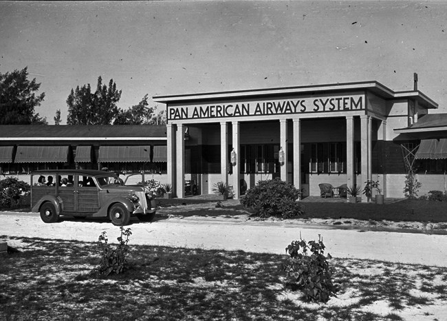 The Pan Am Hotel on Midway, 1930s. Pan Am used similar “woody” style cars at all of their Pacific hotels. Courtesy Pan Am Historical Foundation / paa.org. Black and white photo of a one story building. The center entrance has “Pan American Airways System” over it. Several people sit inside a vehicle driving on a shell-covered roadway. There are small plants and scrub grass.