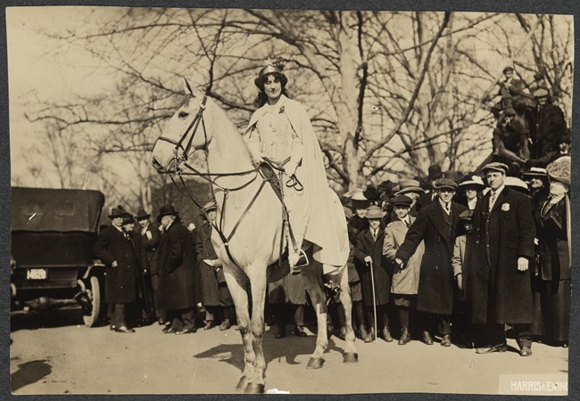 Inez Milholland March 3, 1913 Photograph of Inez Milholland, full-length, facing forward, wearing white robes and a crown, astride a white horse in front of a crowd on a street in Washington, D.C.