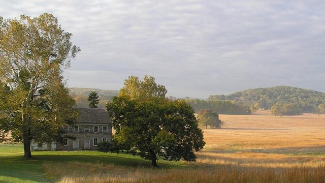 Two-story stone house in field turning brown, tree-covered hills in distance.