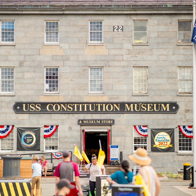 A grey stone 3-stroy rectangular building with a red door in the center and museum sign above.