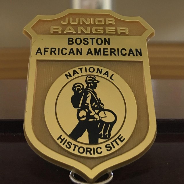 A matte gold ranger bade that has a silhouette of a drummer soldier marching in the center.