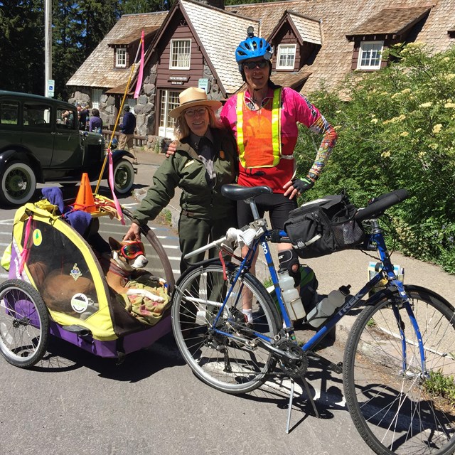 man and ranger behind a blue bicycle with a cart attached and a dog wearing goggles inside