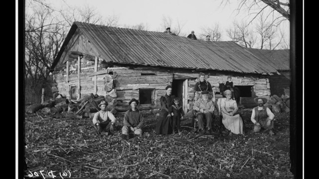 Black and white photo, white family outside log home.