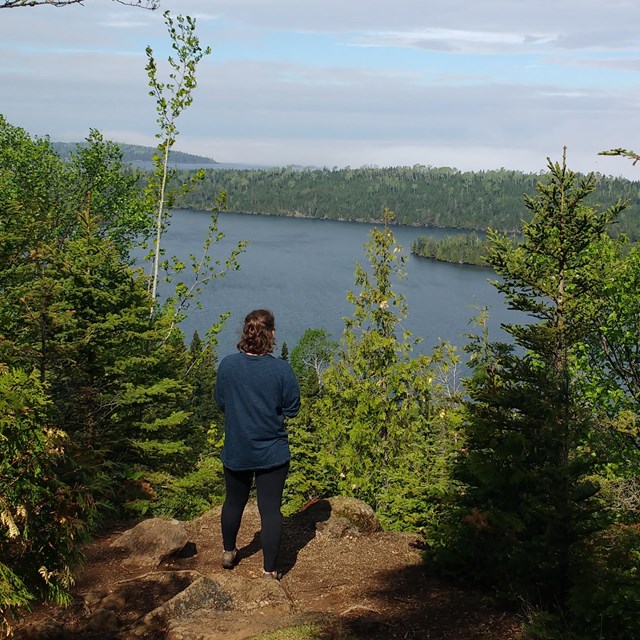 A person's back is to the camera on a high cliff as they look down at a lake and rocks below.