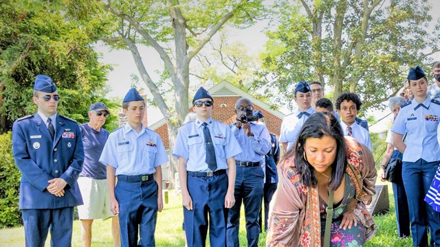 Color photo of a woman placing flowers at a gravestone, while many others people look on
