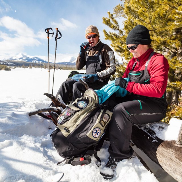 A pair of skiers on change layers and eat while out for a ski.