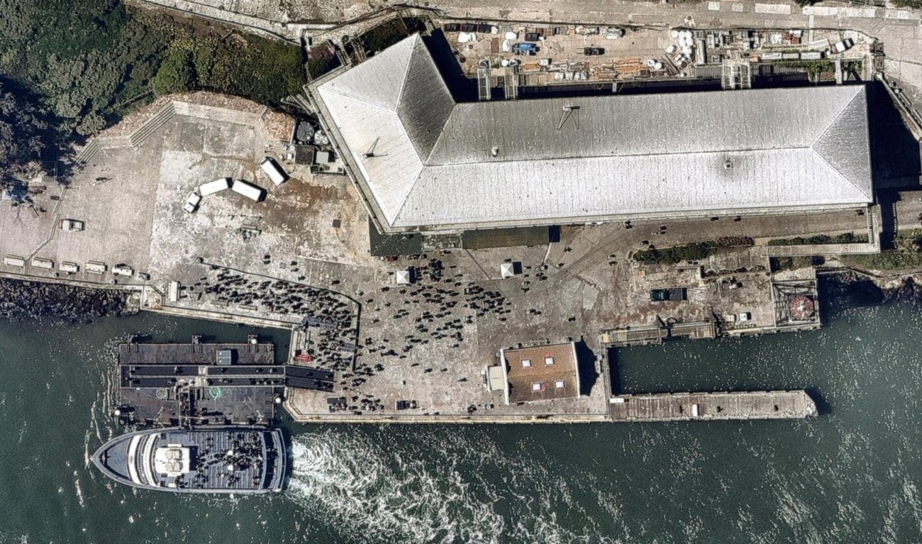 Alcatraz_ConcreteWharf_FingerPier_AerialView Color photo aerial view of Alcatraz concrete wharf with white ferry docked at lower right of picture. Large grey roof of building along top of photo. Dark green water of San Francisco Bay visible at bottom of photo.