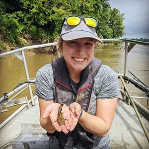 Alexis VandenBerg A scientist in a boat on a river holding a small fish in her hands smiling at the camera.