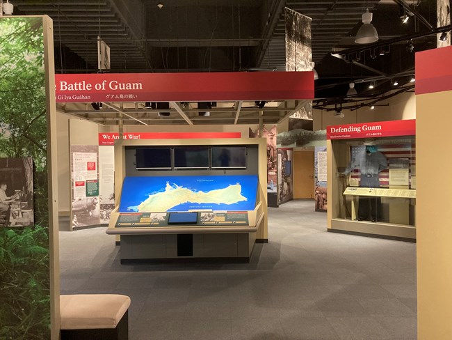 A museum exhibit on the Battle of Guam. A large interactive map display is directly in front of the entrance. To the fight is an exhibit case labelled “Defending Guam” displaying a uniform. Behind the map is a display labelled “We Are at War!”