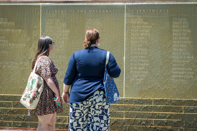 Two women stand and read the names of the signers of the Declaration of Sentiments inscribed on a stone wall; water flows down the wall and runs over the names.